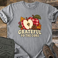 Grateful to the Core Comfort Colors T-shirt in Grey, laid flat. A red gingham apple and an apple half with seeds sit with a pinecone, autumn leaves and berries above the words Grateful to the Core. Sleeves rolled, it lies on weathered wood by olive pants and brown boots.