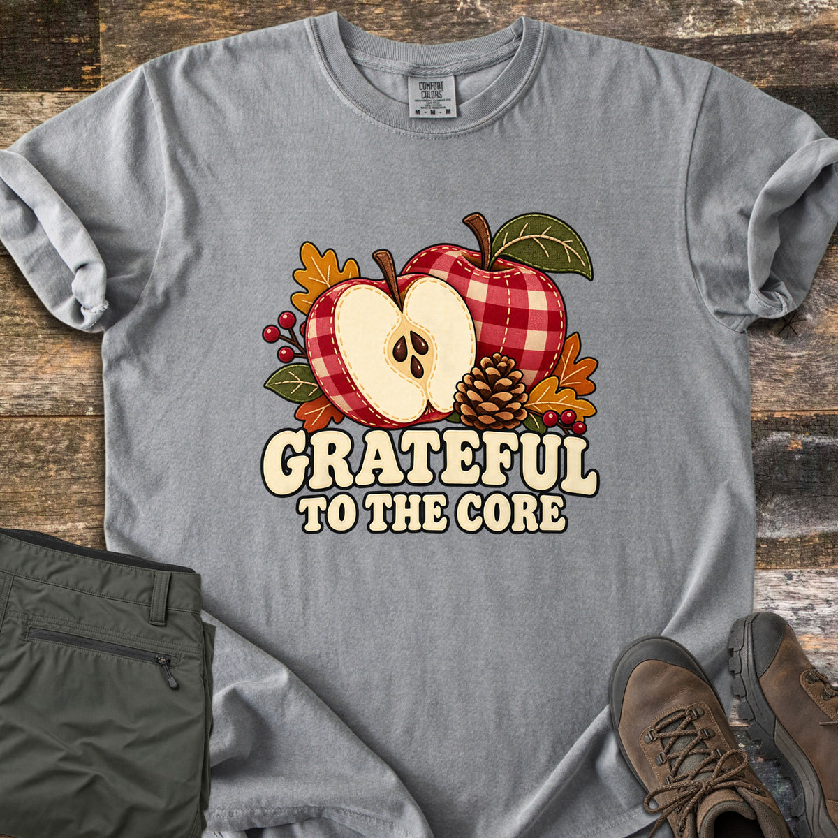 Grateful to the Core Comfort Colors T-shirt in Grey, laid flat. A red gingham apple and an apple half with seeds sit with a pinecone, autumn leaves and berries above the words Grateful to the Core. Sleeves rolled, it lies on weathered wood by olive pants and brown boots.