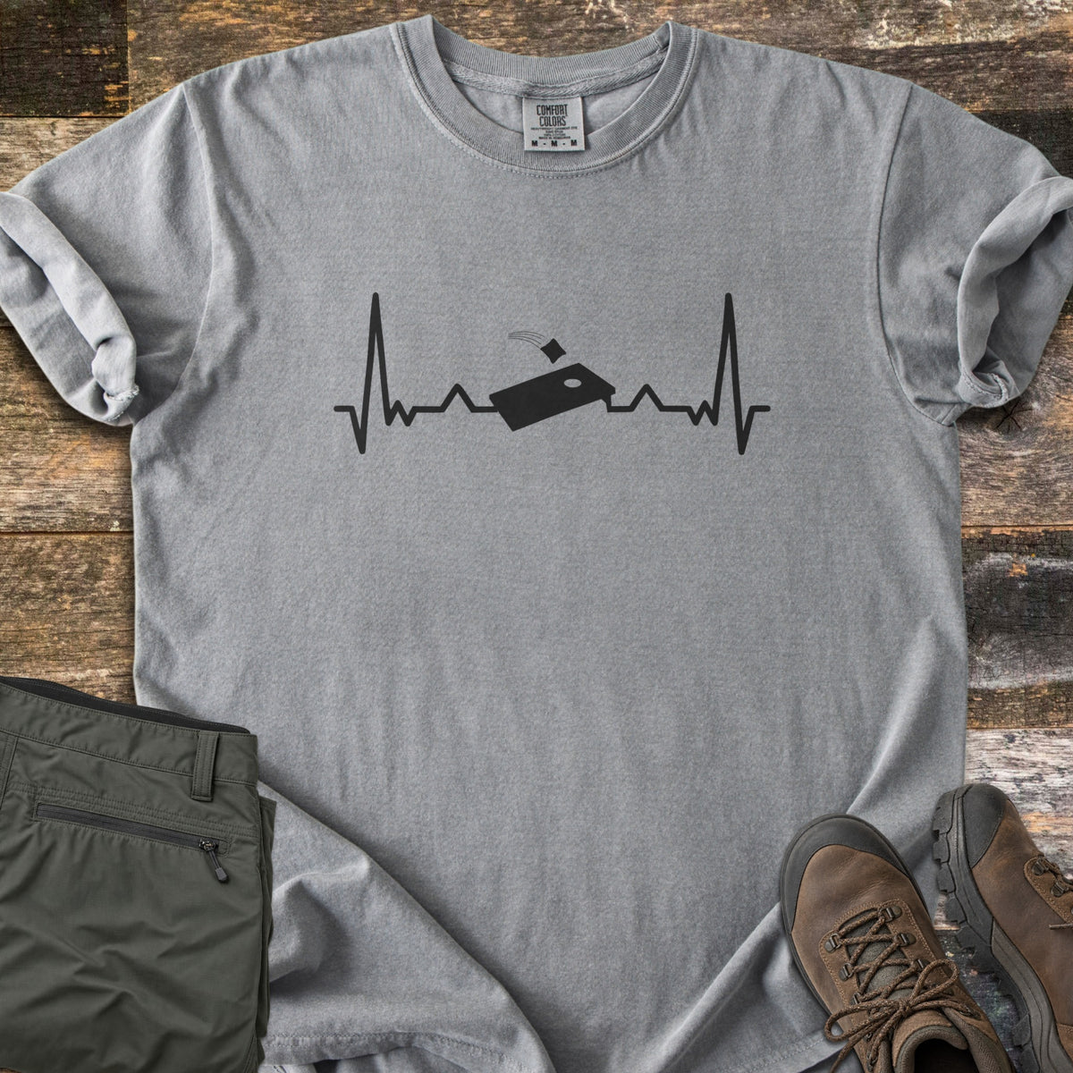 Corn Hole Comfort Colors T-shirt in Grey, laid flat. A black heartbeat line runs across the chest with a cornhole board at its center and a bean bag flying toward the hole. Olive pants and brown hiking boots sit beside it on weathered wood planks.