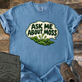 Ask Me About Moss Comfort Colors T-shirt in Blue Jean, laid flat. A cream speech bubble reads Ask Me About Moss in dark green letters above a mound of green moss with three spore stalks. Sleeves rolled, on weathered wood with folded olive hiking pants and brown hiking boots.