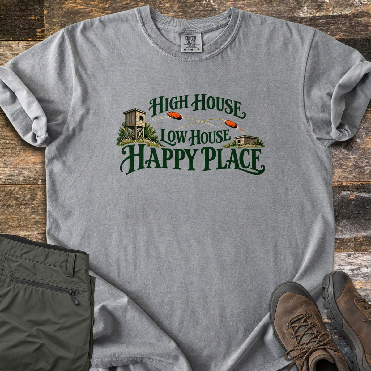High House Low House Happy Place Comfort Colors T-shirt in Grey, laid flat. Green lettering reads High House Low House Happy Place around two skeet houses with orange clay targets flying between them. Olive cargo pants and brown hiking boots lie beside it on weathered wood planks.