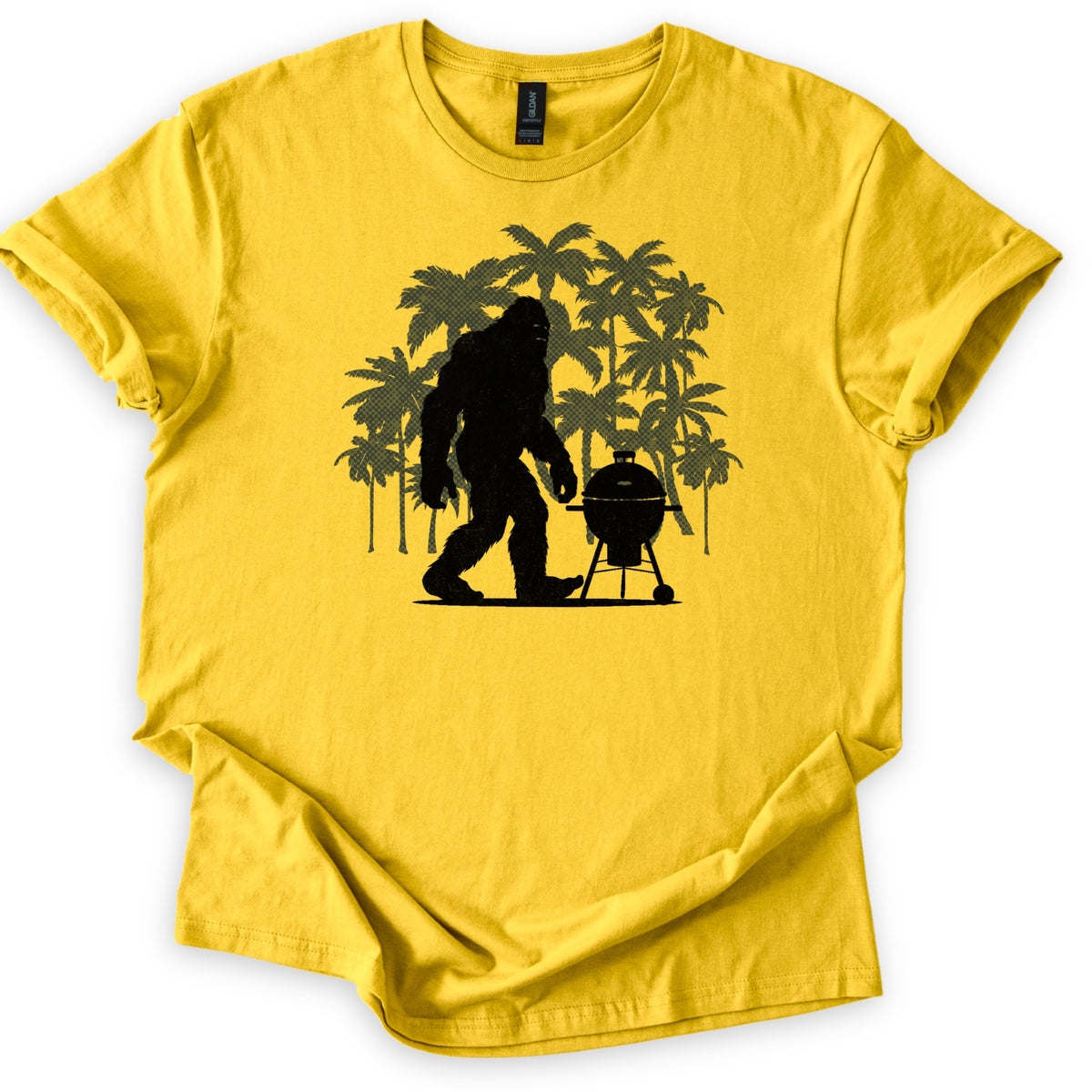 Squatch BBQ Smoker Comfort Colors T-shirt in Daisy, laid flat. A black silhouette of Bigfoot walks beside a kettle-style grill on wheeled legs, set against a cluster of olive green palm trees. No words are printed. The shirt lies on a white surface with sleeves cuffed.