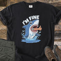 Shark I'm Fine Comfort Colors T-shirt in Black, laid flat. A cartoon shark leaps from splashing water with a human arm giving a thumbs up sticking out of its mouth, under the words I'M FINE. Olive cargo pants and brown hiking boots sit beside it on weathered wood.