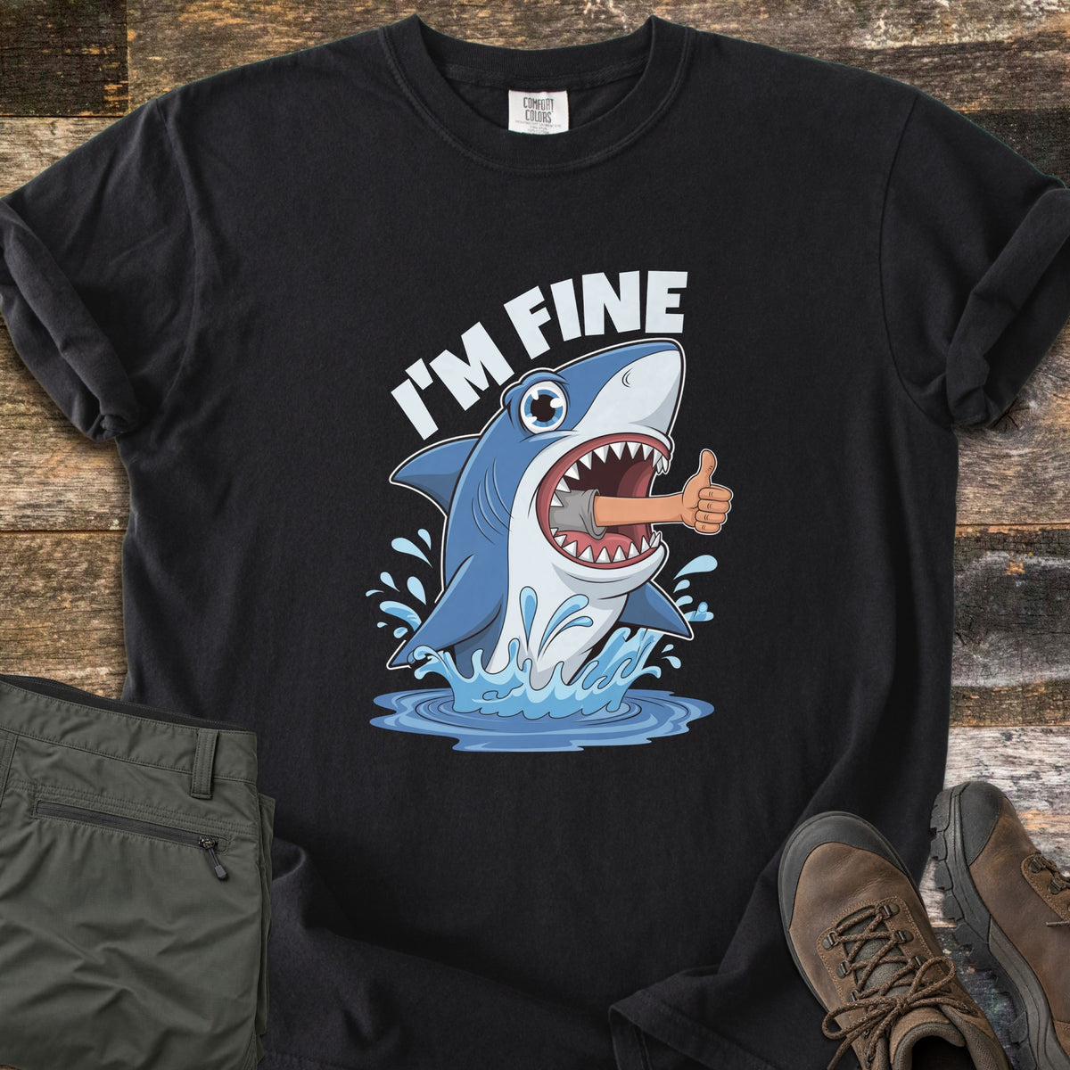 Shark I'm Fine Comfort Colors T-shirt in Black, laid flat. A cartoon shark leaps from splashing water with a human arm giving a thumbs up sticking out of its mouth, under the words I'M FINE. Olive cargo pants and brown hiking boots sit beside it on weathered wood.