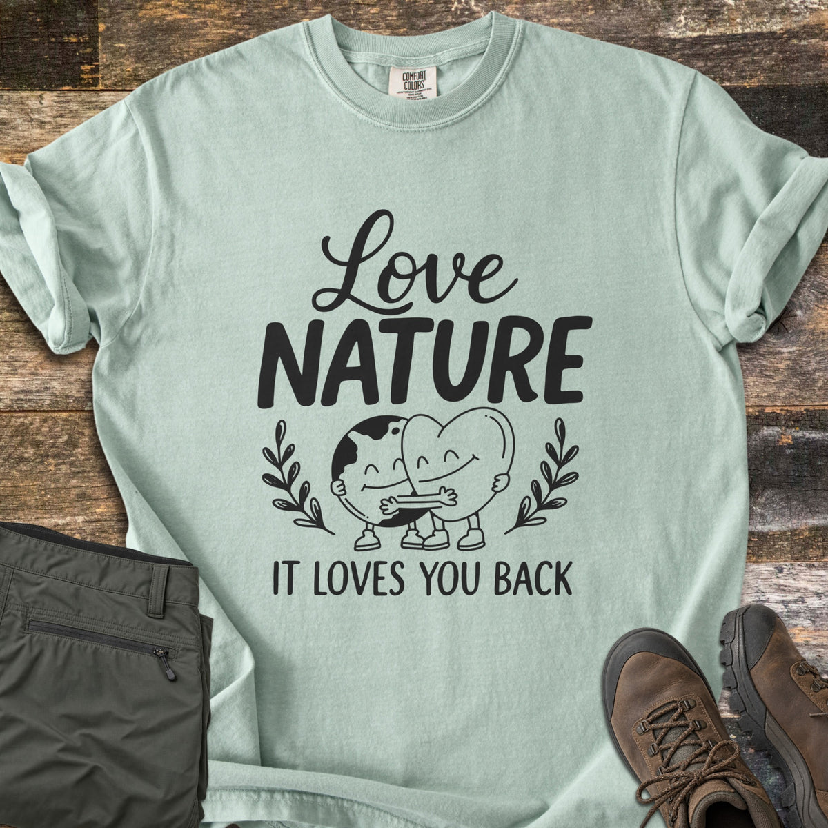 Nature Loves You Back Comfort Colors T-shirt in Bay, laid flat. Black print reads Love Nature above a smiling cartoon Earth and heart hugging between leafy sprigs, with It Loves You Back below. It lies on weathered wood beside olive hiking pants and brown boots.