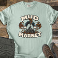 Mud Magnet Comfort Colors T-shirt in Bay, laid flat. The words MUD MAGNET arch around a horseshoe magnet pulling mud splashes toward an ATV rider churning through mud. Shown on a wood floor with folded olive pants and brown hiking boots.