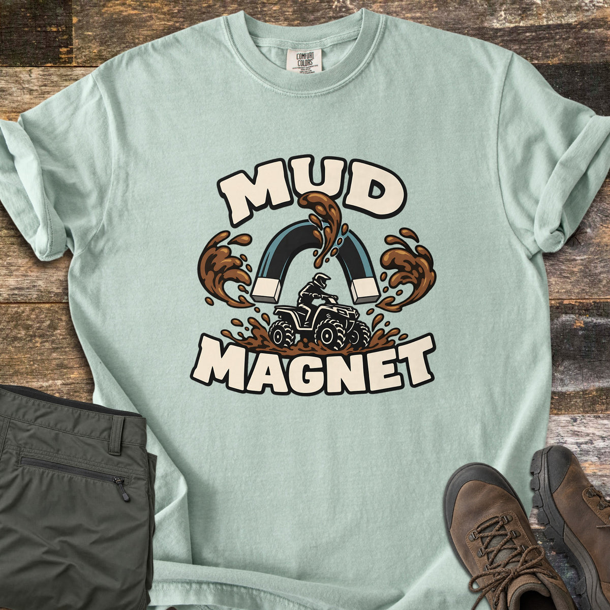 Mud Magnet Comfort Colors T-shirt in Bay, laid flat. The words MUD MAGNET arch around a horseshoe magnet pulling mud splashes toward an ATV rider churning through mud. Shown on a wood floor with folded olive pants and brown hiking boots.