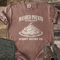 Mashed Potato Alpine Club Comfort Colors T-shirt in Espresso, laid flat. An etched drawing of a mashed potato mountain on a plate, with tiny climbers and a flag on a butter pat summit, under Mashed Potato Alpine Club and above Summit Before Pie. Green hiking pants and boots sit on wood.
