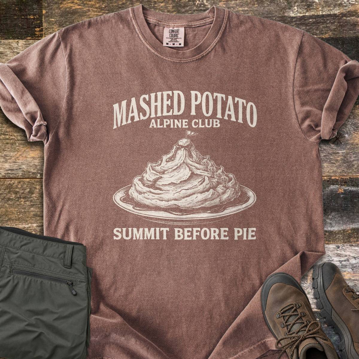 Mashed Potato Alpine Club Comfort Colors T-shirt in Espresso, laid flat. An etched drawing of a mashed potato mountain on a plate, with tiny climbers and a flag on a butter pat summit, under Mashed Potato Alpine Club and above Summit Before Pie. Green hiking pants and boots sit on wood.