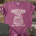 Backyard Smokehouse Comfort Colors T-shirt in Berry, laid flat. Cream print of a smoker grill with a chicken inside and a folding chair beside it, under the words Backyard Smokehouse and above Ready When It's Ready. Olive pants and brown boots lie on weathered wood.