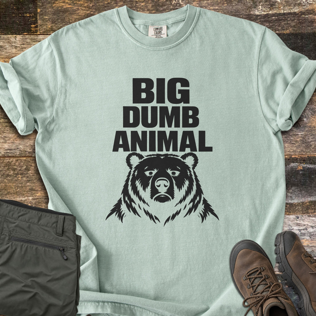 Big Dumb Animal Comfort Colors T-shirt in Bay, laid flat. Bold black block letters read Big Dumb Animal above a grumpy-faced bear head. The shirt, sleeves rolled, lies on weathered wood planks beside folded olive hiking pants and brown hiking boots.