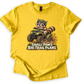 Small Paws Big Trail Plans Comfort Colors T-shirt in Daisy, laid flat. A cartoon raccoon in a green helmet rides a muddy green ATV, kicking up dirt beside pine trees, above the words Small Paws Big Trail Plans. The shirt lies on a white background with cuffed sleeves.