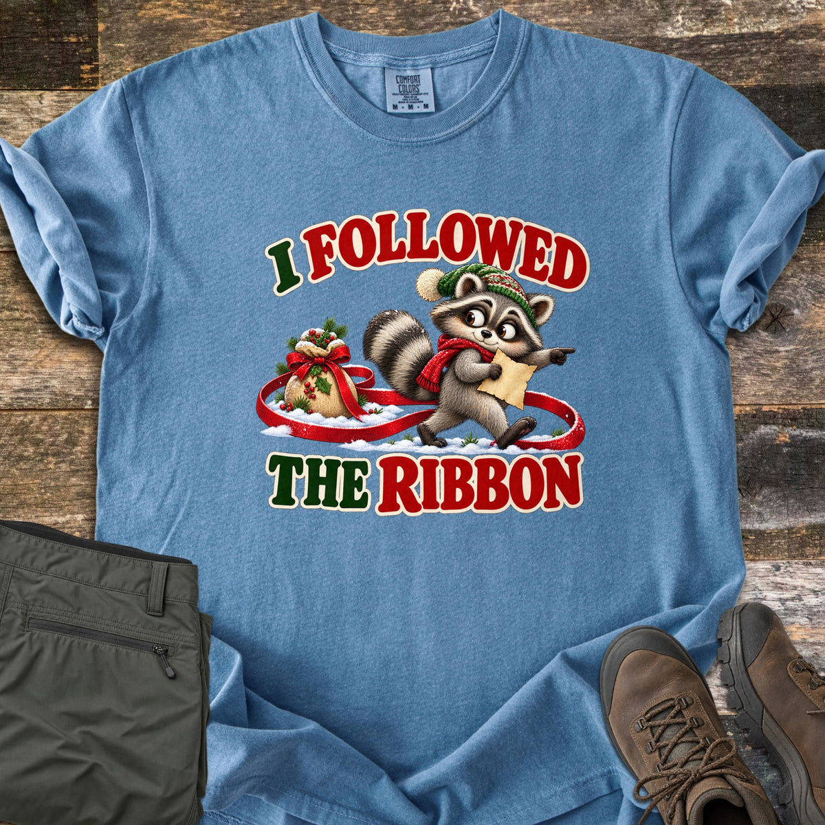 I Followed The Ribbon Comfort Colors T-shirt in Blue Jean, laid flat. A cartoon raccoon in a green knit hat and red scarf holds a map and points ahead, walking along a red ribbon from a gift sack in snow, between the words I Followed The Ribbon. Olive pants and brown boots lie on a wood floor.