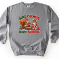 Some Assembly. Mostly Squirrels. Comfort Colors crewneck sweatshirt in Grey, laid flat. An orange squirrel in a scarf holds a red ornament on a snowy stump beside a bunny wrapped in red ribbon, between the words Some Assembly. Mostly Squirrels. Laid on a white background.