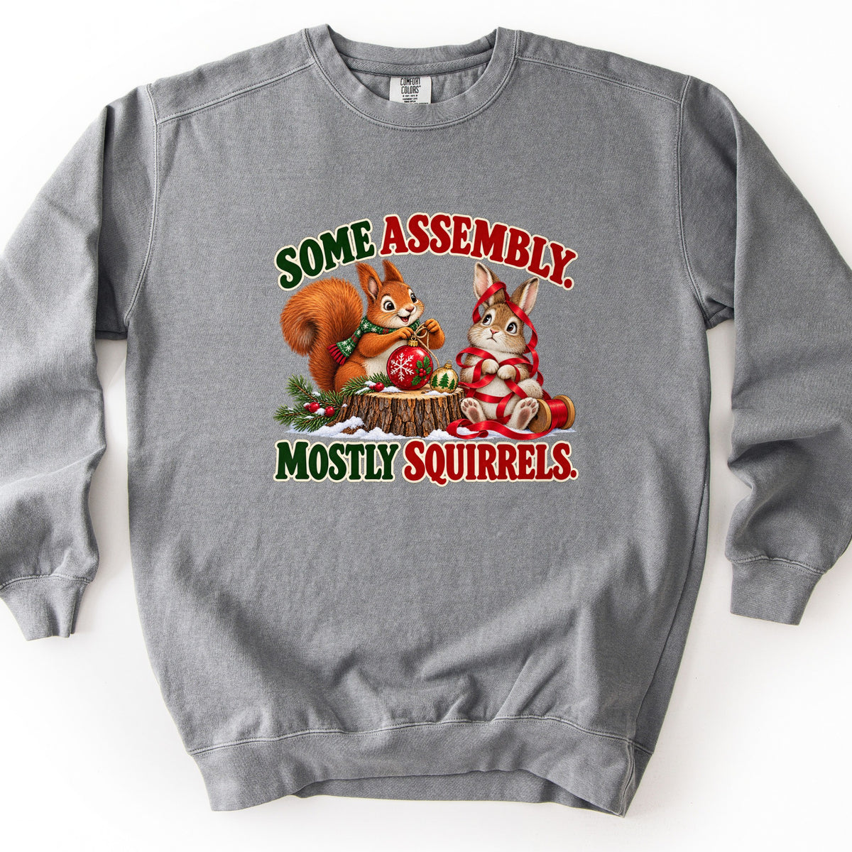 Some Assembly. Mostly Squirrels. Comfort Colors crewneck sweatshirt in Grey, laid flat. An orange squirrel in a scarf holds a red ornament on a snowy stump beside a bunny wrapped in red ribbon, between the words Some Assembly. Mostly Squirrels. Laid on a white background.
