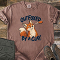 Outfoxed by a Clay Comfort Colors T-shirt in Espresso, laid flat. A cartoon red fox glances sideways at an orange clay target flying past, between the words Outfoxed By A Clay in navy brush letters. Sleeves rolled on rustic wood with olive pants and brown hiking boots.