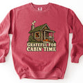 Grateful For Cabin Time Comfort Colors crewneck sweatshirt in Crimson, laid flat. A log cabin with a green roof, smoking stone chimney, glowing window, pie on the porch rail and a pumpkin by the steps, above the words Grateful For Cabin Time. The sweatshirt lies on a white surface.
