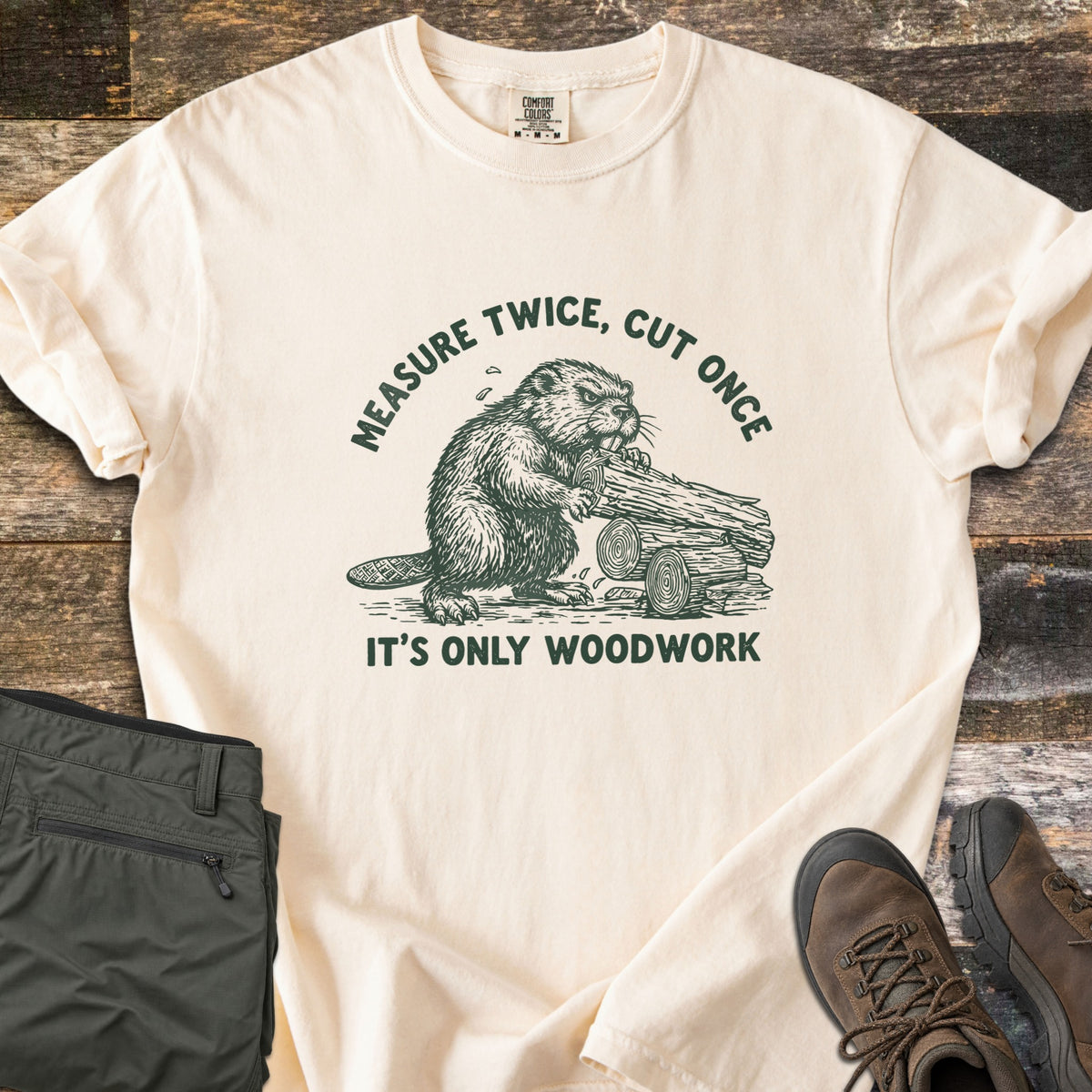 Measure Twice Beaver Comfort Colors T-shirt in Ivory, laid flat. A green sketch of a beaver gnawing a pile of logs, with Measure Twice, Cut Once arched above and It's Only Woodwork below. Folded olive cargo pants and brown hiking boots sit on a weathered wood surface.