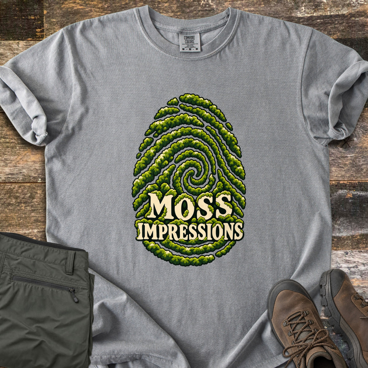 Moss Impressions Comfort Colors T-shirt in Grey, laid flat. A fingerprint made of green moss ridges with the words Moss Impressions in cream letters. The shirt lies on weathered wood with folded olive hiking pants and brown hiking boots.