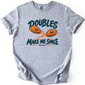 Doubles Make Me Smile Comfort Colors T-shirt in Sport Grey, laid flat. Two smiling orange clay targets with motion streaks fly between the words Doubles and Make Me Smile in dark teal, with Skeet in small letters below. The shirt has rolled sleeves on a white background.
