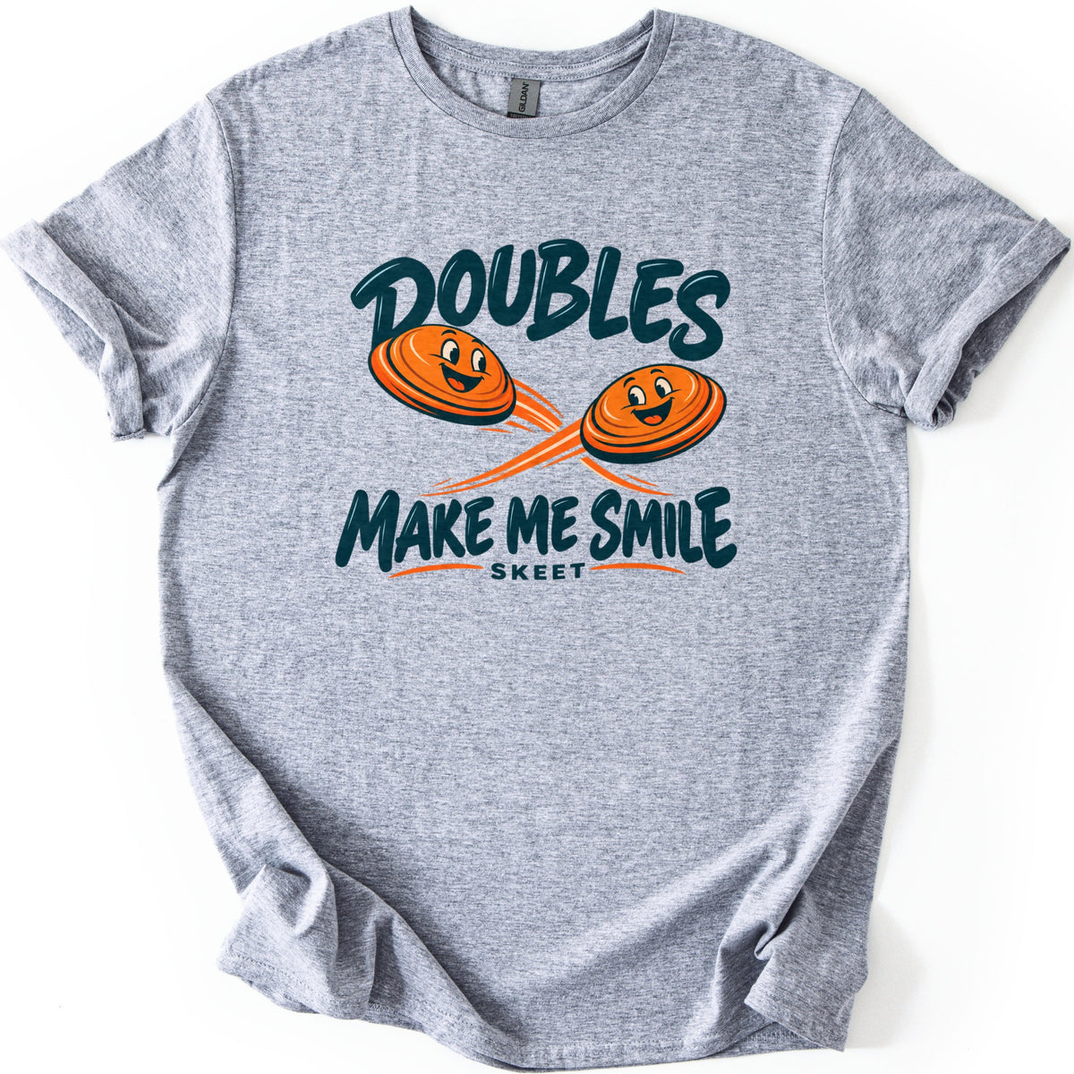 Doubles Make Me Smile Comfort Colors T-shirt in Sport Grey, laid flat. Two smiling orange clay targets with motion streaks fly between the words Doubles and Make Me Smile in dark teal, with Skeet in small letters below. The shirt has rolled sleeves on a white background.