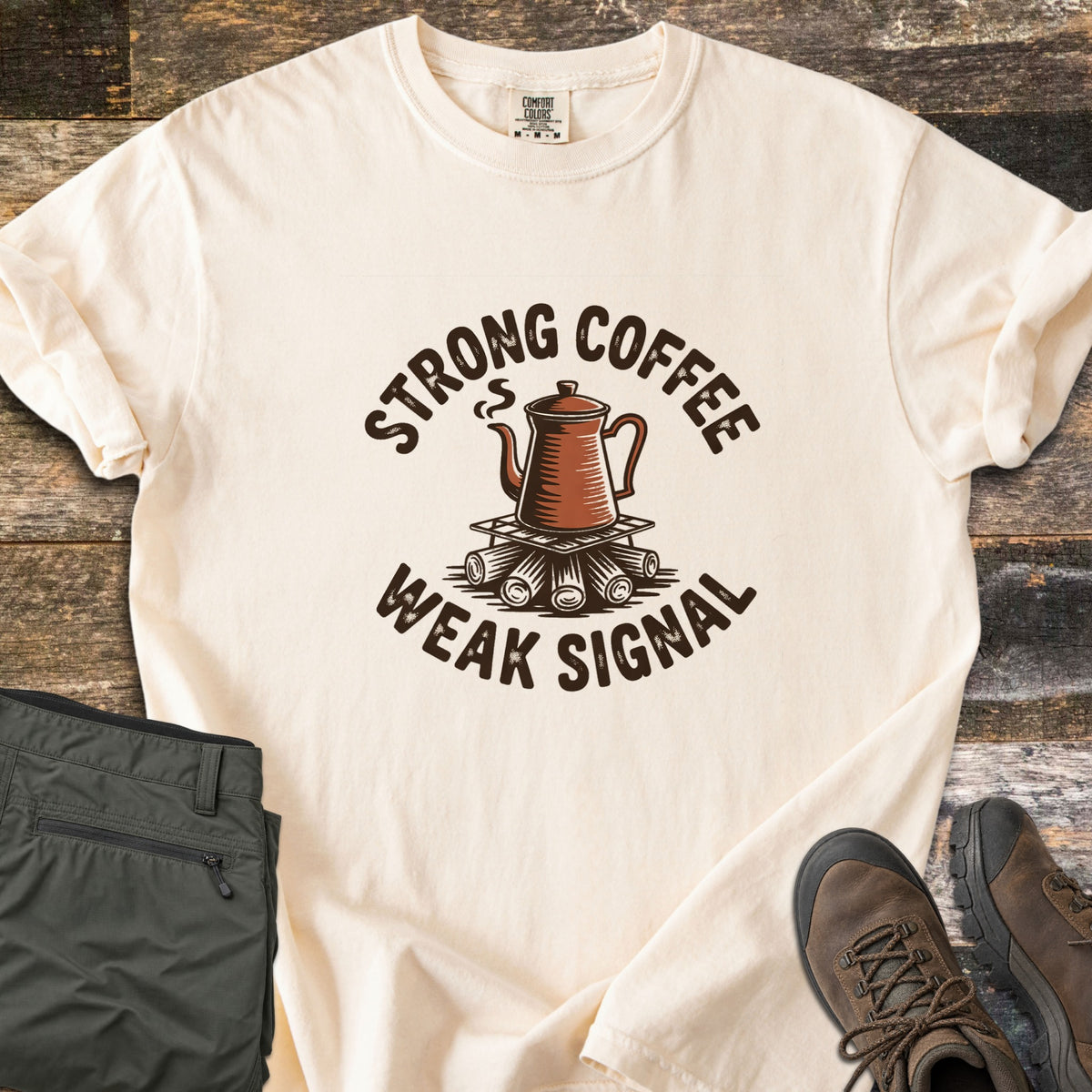 Strong Coffee Weak Signal Comfort Colors T-shirt in Ivory, laid flat. A steaming copper coffee pot sits on a grate over campfire logs, arched by the words Strong Coffee above and Weak Signal below. Folded olive hiking pants and brown hiking boots lie on weathered wood planks.