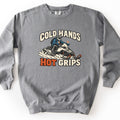 Cold Hands Hot Grips Comfort Colors crewneck sweatshirt in Grey, laid flat. A rider in a blue jacket and white helmet drives a snowmobile through spraying snow, small orange flames rising from his handlebar grips, between the words Cold Hands and Hot Grips, with Hot in orange.