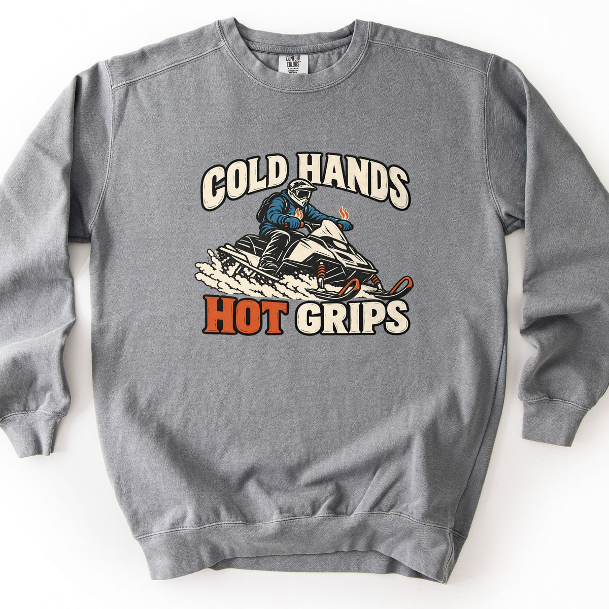 Cold Hands Hot Grips Comfort Colors crewneck sweatshirt in Grey, laid flat. A rider in a blue jacket and white helmet drives a snowmobile through spraying snow, small orange flames rising from his handlebar grips, between the words Cold Hands and Hot Grips, with Hot in orange.