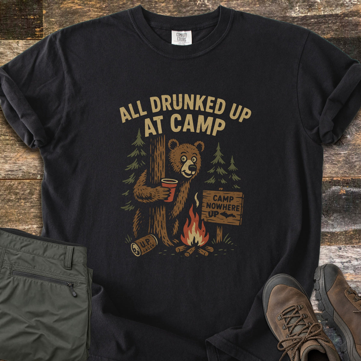 All Drunked Up At Camp Comfort Colors T-shirt in Black, laid flat. A wide-eyed brown bear hugs a tree holding a red cup by a campfire, a U.P. Lager can and a sign reading Camp Nowhere UP, under the words All Drunked Up At Camp. Olive pants and hiking boots lie on weathered wood.