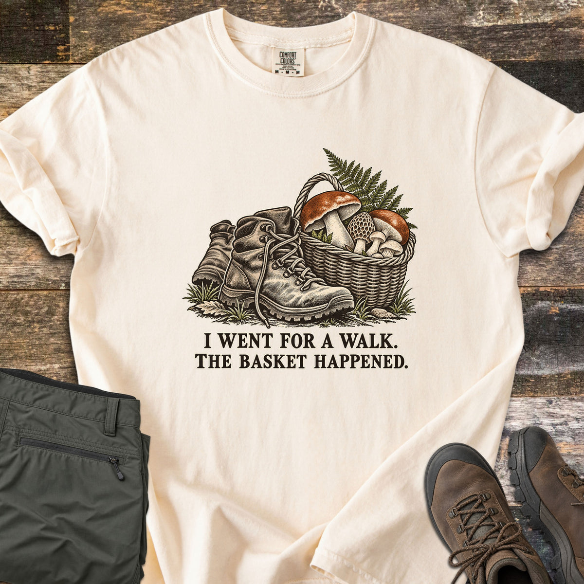 I Went for a Walk. The Basket Happened Comfort Colors T-shirt in Ivory, laid flat. A vintage-style illustration of worn hiking boots beside a wicker basket of mushrooms and fern, above the words I Went for a Walk. The Basket Happened. On rustic wood with olive cargo pants and brown hiking boots.