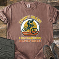 I Don't Crash I Do Random Gravity Checks Comfort Colors T-shirt in Espresso, laid flat. A mountain biker in a full-face helmet rides over orange hills and pines before a yellow sun, between the words I Don't Crash and I Do Random Gravity Checks. Olive pants and brown hiking boots lie on weathered wood.