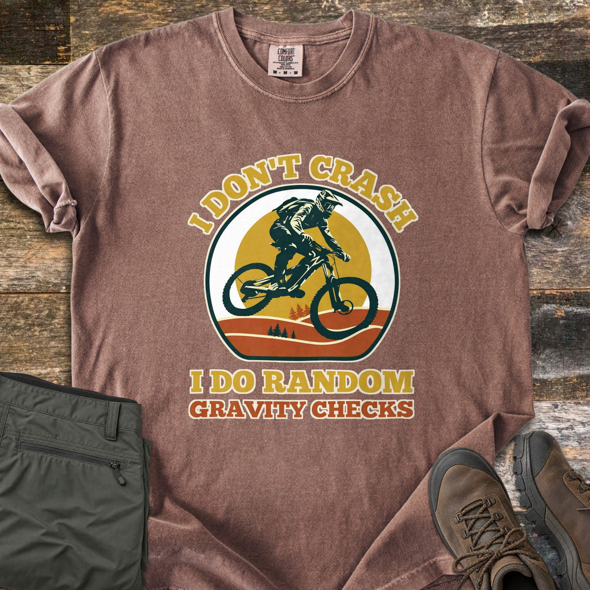 I Don't Crash I Do Random Gravity Checks Comfort Colors T-shirt in Espresso, laid flat. A mountain biker in a full-face helmet rides over orange hills and pines before a yellow sun, between the words I Don't Crash and I Do Random Gravity Checks. Olive pants and brown hiking boots lie on weathered wood.