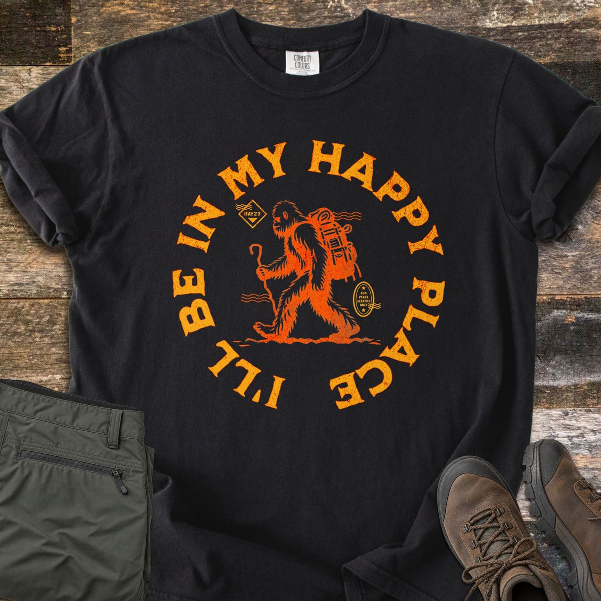 Happy Place Hiking Squatch Comfort Colors T-shirt in Black, laid flat. Orange distressed art of Bigfoot hiking with a walking stick and a large backpack, circled by the words I'll Be In My Happy Place. Folded olive hiking pants and brown hiking boots lie beside it on weathered wood.