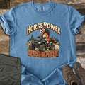 Horsepower Upgrade Comfort Colors T-shirt in Blue Jean, laid flat. A cartoon horse in a green helmet rides a muddy green ATV past pine trees, between the words HorsePower and UPGRADE. Olive cargo pants and brown hiking boots lie beside it on weathered wood.