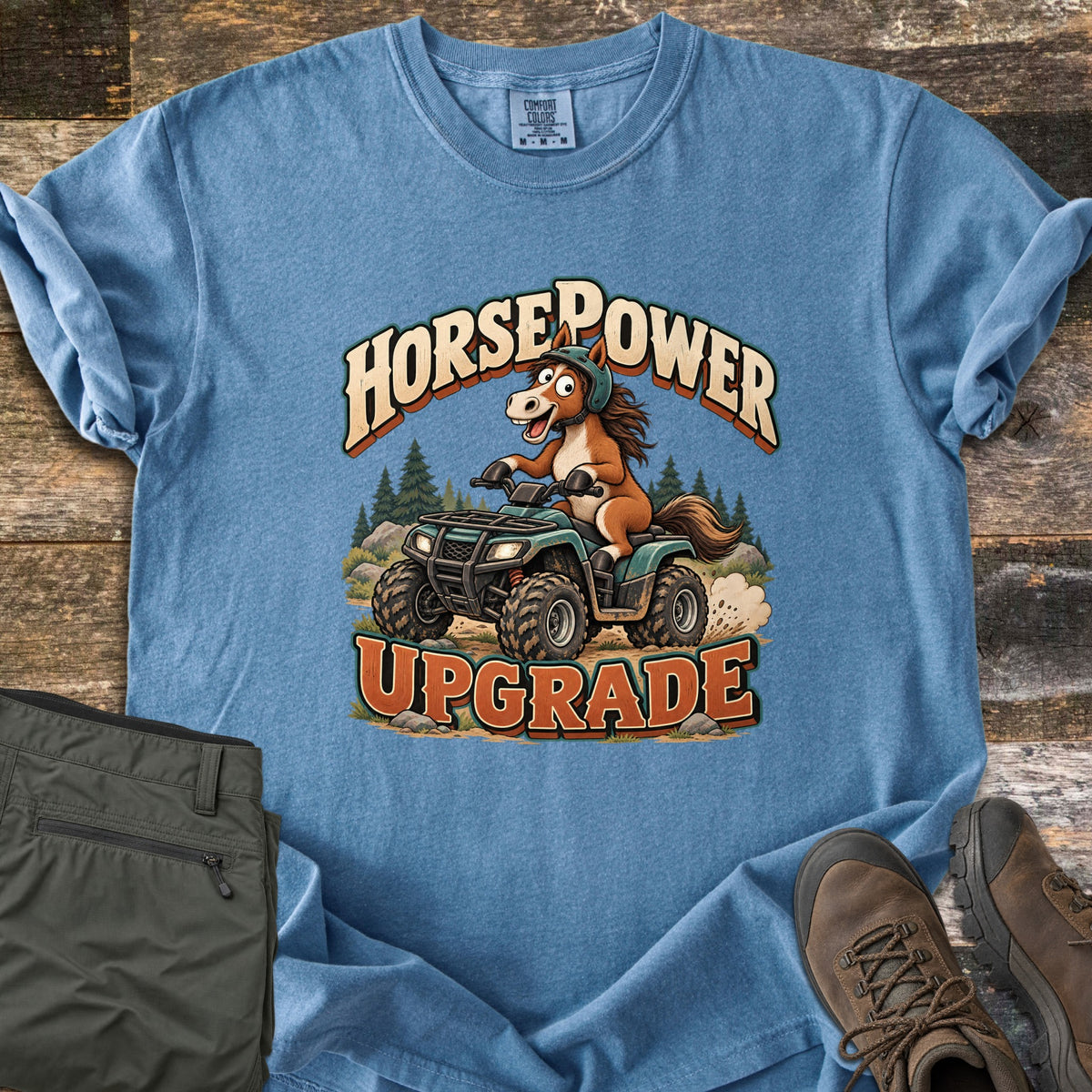 Horsepower Upgrade Comfort Colors T-shirt in Blue Jean, laid flat. A cartoon horse in a green helmet rides a muddy green ATV past pine trees, between the words HorsePower and UPGRADE. Olive cargo pants and brown hiking boots lie beside it on weathered wood.