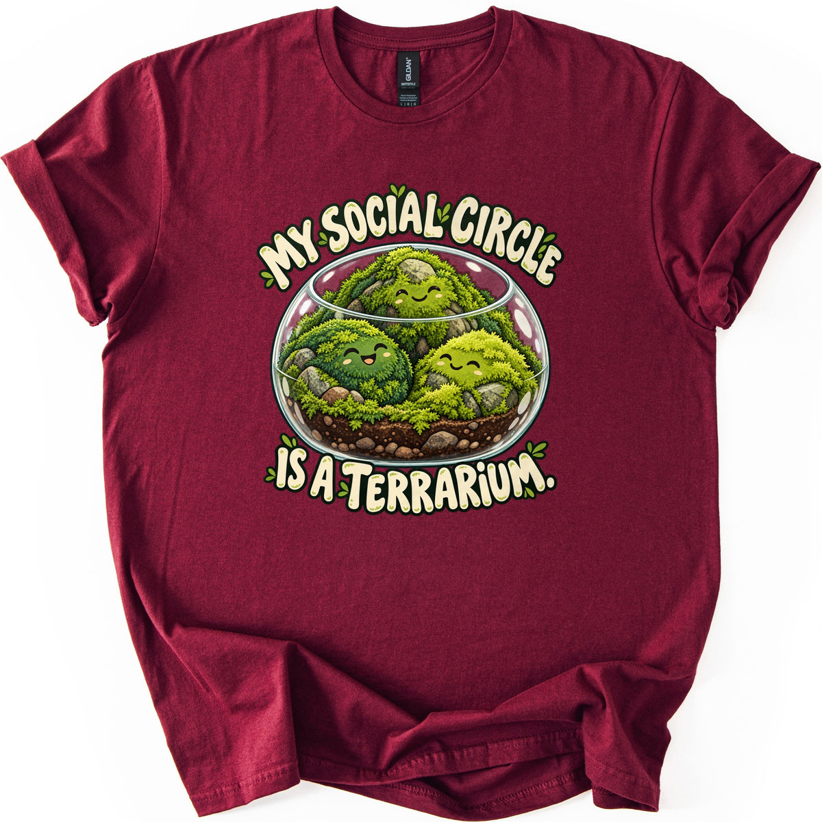 My Social Circle Is a Terrarium Comfort Colors T-shirt in Maroon, laid flat. Three smiling moss balls sit among stones and soil in a round glass terrarium, with the words My Social Circle above and Is a Terrarium. below in cream letters. Sleeves are cuffed on a white background.