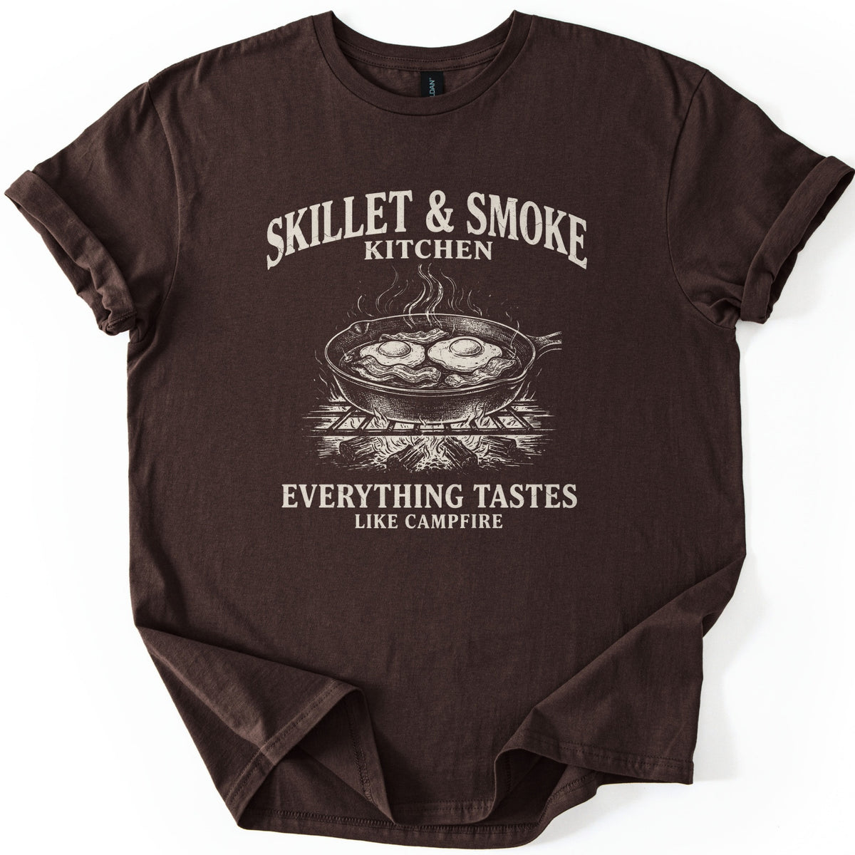 Skillet & Smoke Kitchen Comfort Colors T-shirt in Dark Chocolate, laid flat. A cream print shows a cast iron skillet of fried eggs and bacon steaming on a grate over a campfire, under the words Skillet & Smoke Kitchen and above Everything Tastes Like Campfire. Sleeves cuffed, on white.