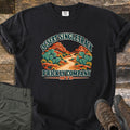 Desert Singletrack Prickly Company Comfort Colors T-shirt in Black, laid flat. A winding dirt trail runs between prickly pear cacti and red rock buttes under the words Desert Singletrack, with Prickly Company MTB below. Olive pants and brown hiking boots lie on weathered wood boards.