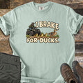 I Brake for Ducks Comfort Colors T-shirt in Bay, laid flat. A rider in a helmet on a green ATV waits as a mother duck leads her ducklings across a dirt path, under the words I Brake For Ducks. Olive pants and brown hiking boots lie beside it on weathered wood planks.
