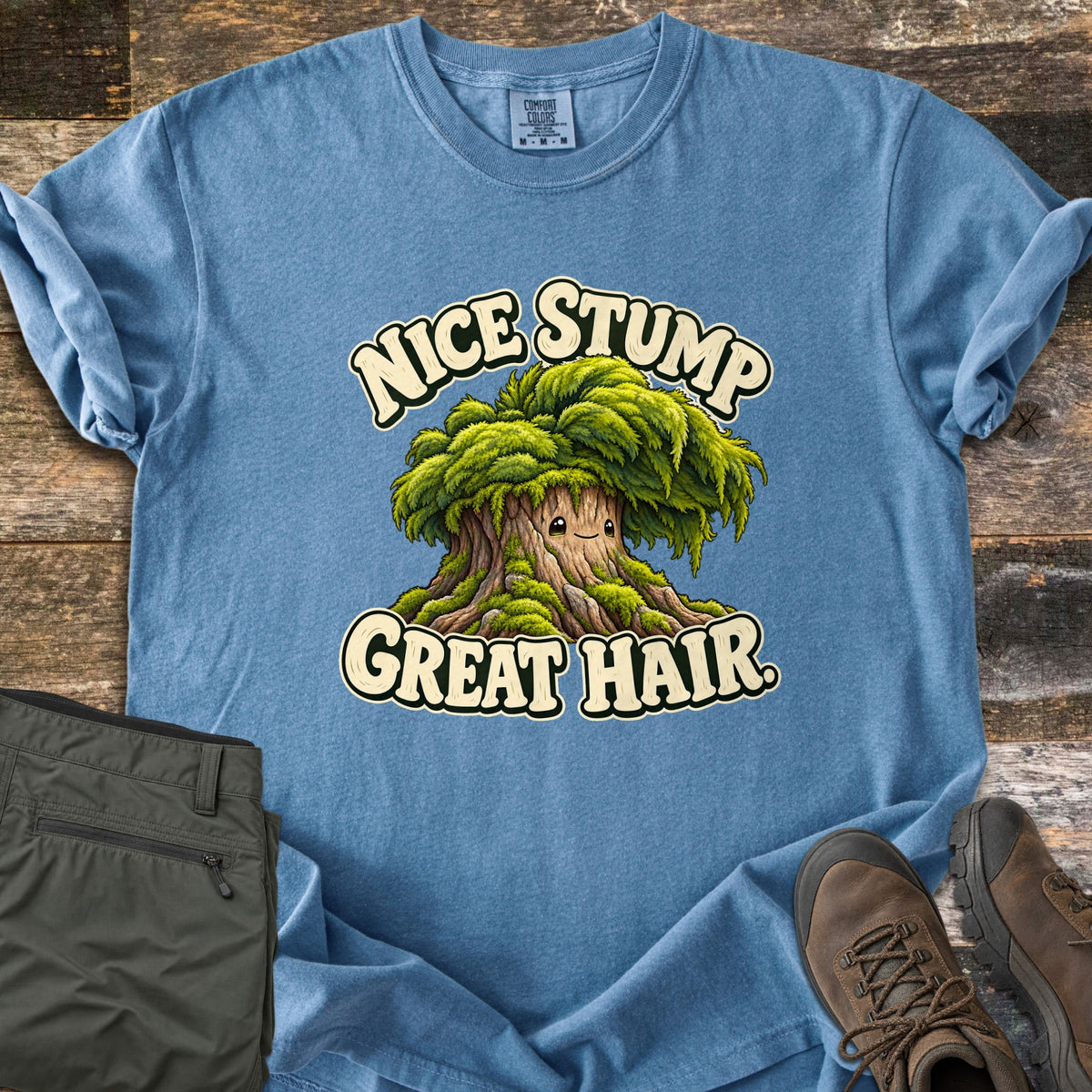 Nice Stump Great Hair Comfort Colors T-shirt in Blue Jean, laid flat. A smiling cartoon tree stump with a shaggy mop of green moss hair sits between the words Nice Stump Great Hair. Sleeves rolled, on weathered wood with folded olive hiking pants and brown hiking boots.