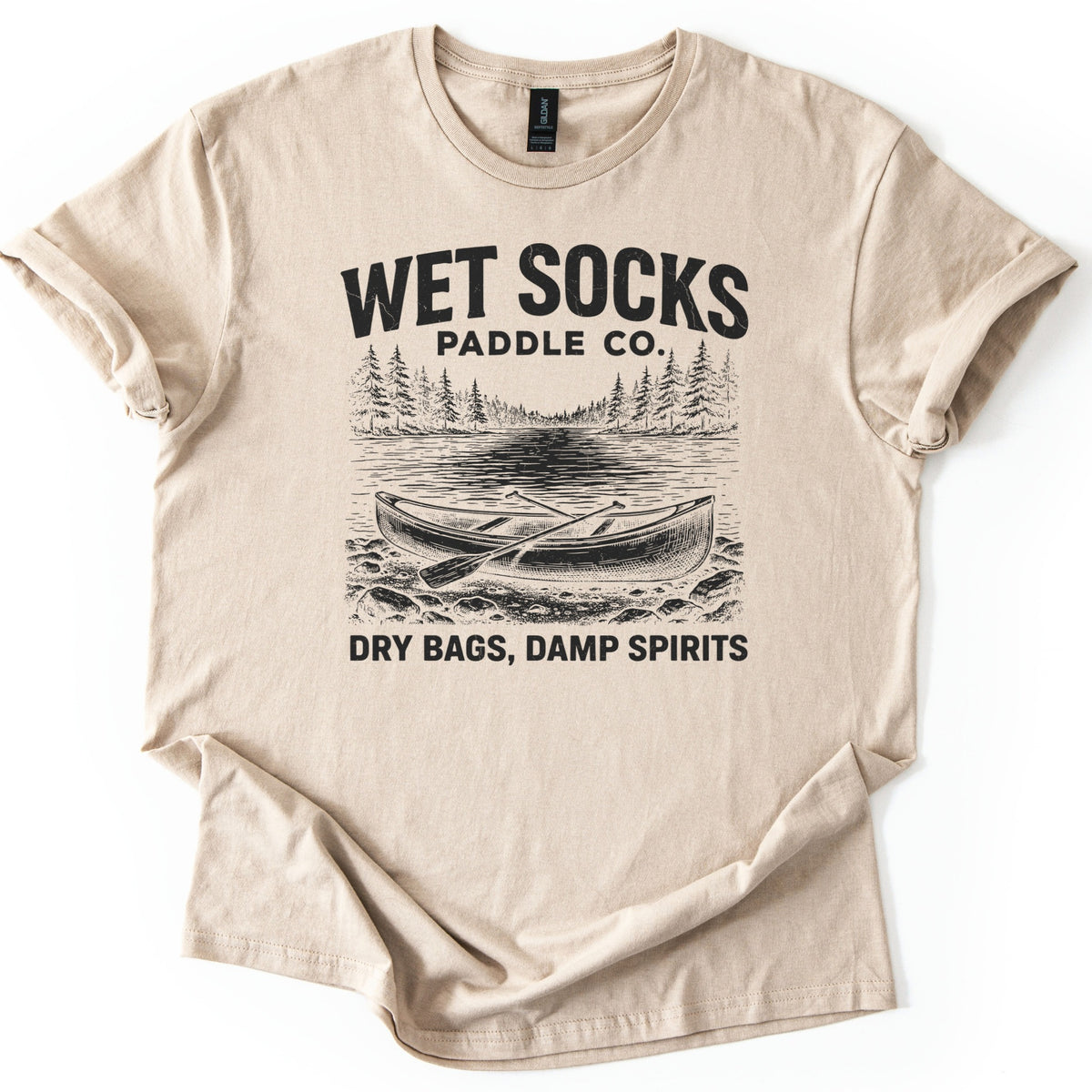Wet Socks Paddle Co Comfort Colors T-shirt in Sand, laid flat. A black etched drawing of a canoe with crossed paddles on a rocky shore, with a lake and pine trees behind, under the words Wet Socks Paddle Co. and above Dry Bags, Damp Spirits. White background.