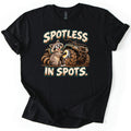 Spotless in Spots Comfort Colors T-shirt in Black, laid flat. A grinning cartoon raccoon scrubs a mud-caked ATV tire with a soapy sponge between the words Spotless In Spots. in mud-spattered cream letters. The shirt has rolled sleeves on a white background.