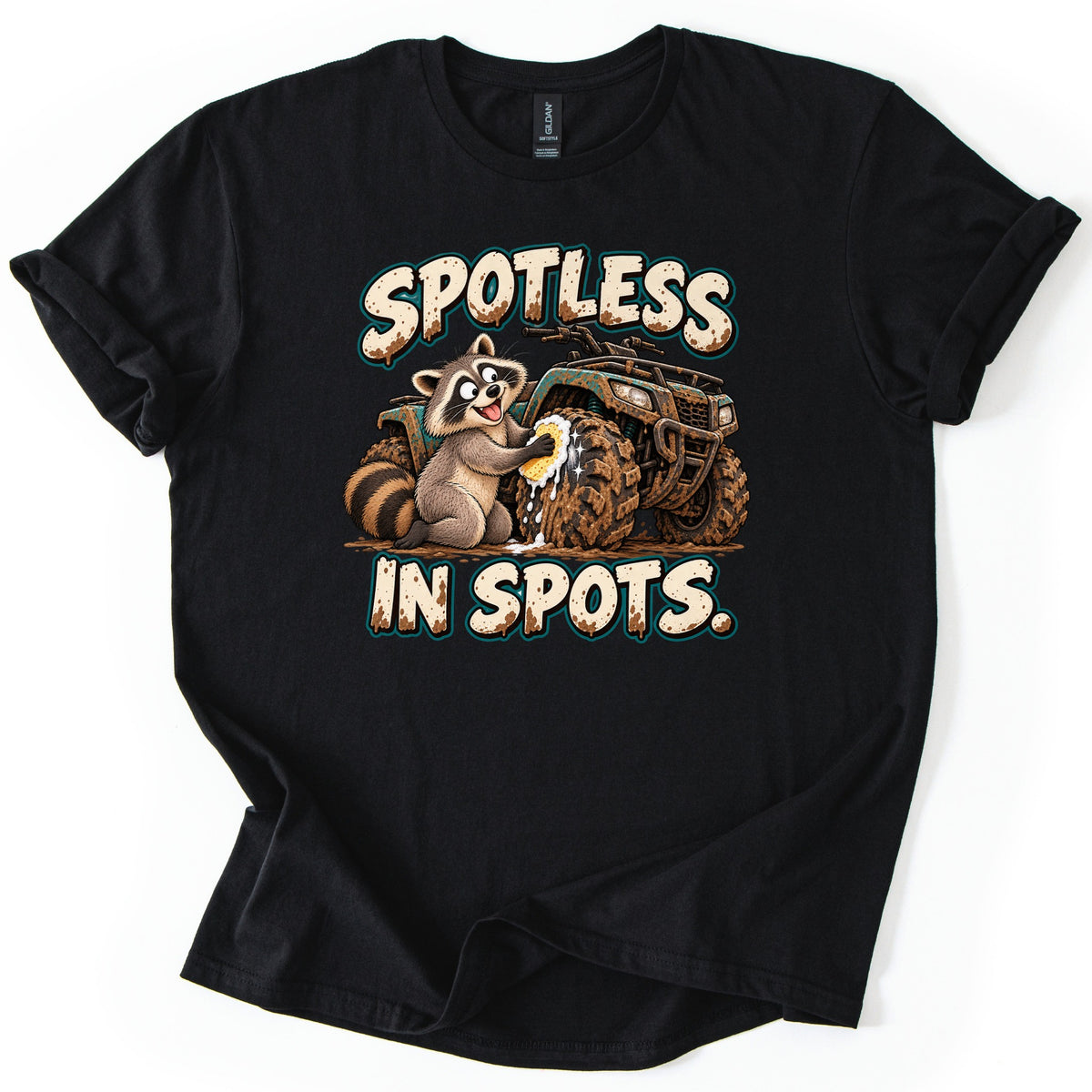 Spotless in Spots Comfort Colors T-shirt in Black, laid flat. A grinning cartoon raccoon scrubs a mud-caked ATV tire with a soapy sponge between the words Spotless In Spots. in mud-spattered cream letters. The shirt has rolled sleeves on a white background.