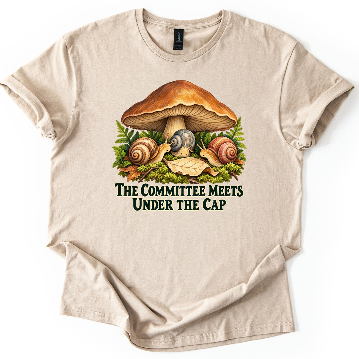 The Committee Meets Under the Cap Comfort Colors T-shirt in Sand, laid flat. A large brown mushroom shelters three snails on moss among ferns and a fallen leaf, above dark green words The Committee Meets Under the Cap. The shirt lies on a white background with rolled sleeves.