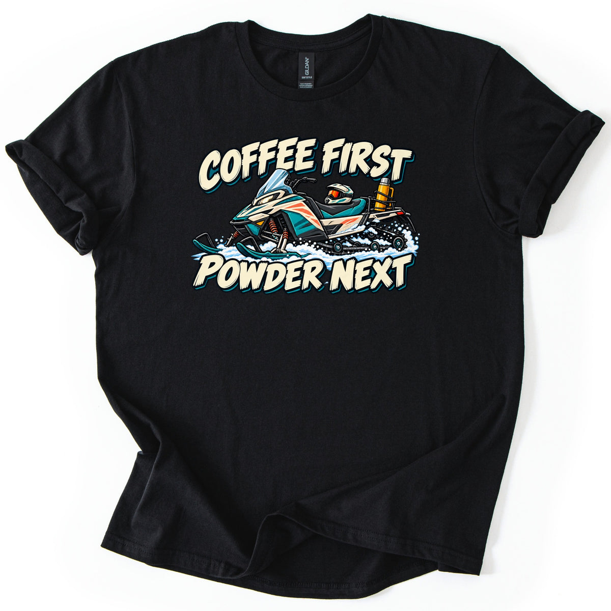 Coffee First Powder Next Comfort Colors T-shirt in Black, laid flat. A teal and cream cartoon snowmobile kicks up snow, with a helmet on the seat and a thermos on the back, between the words Coffee First Powder Next. Sleeves are rolled on a white background.