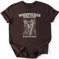 Woodpecker Demolition Comfort Colors T-shirt in Dark Chocolate, laid flat. A cream engraving-style woodpecker pecks splinters from a broken tree trunk under the words WOODPECKER DEMOLITION, with NO JOB TOO SMALL below. The sleeves are cuffed on a white background.