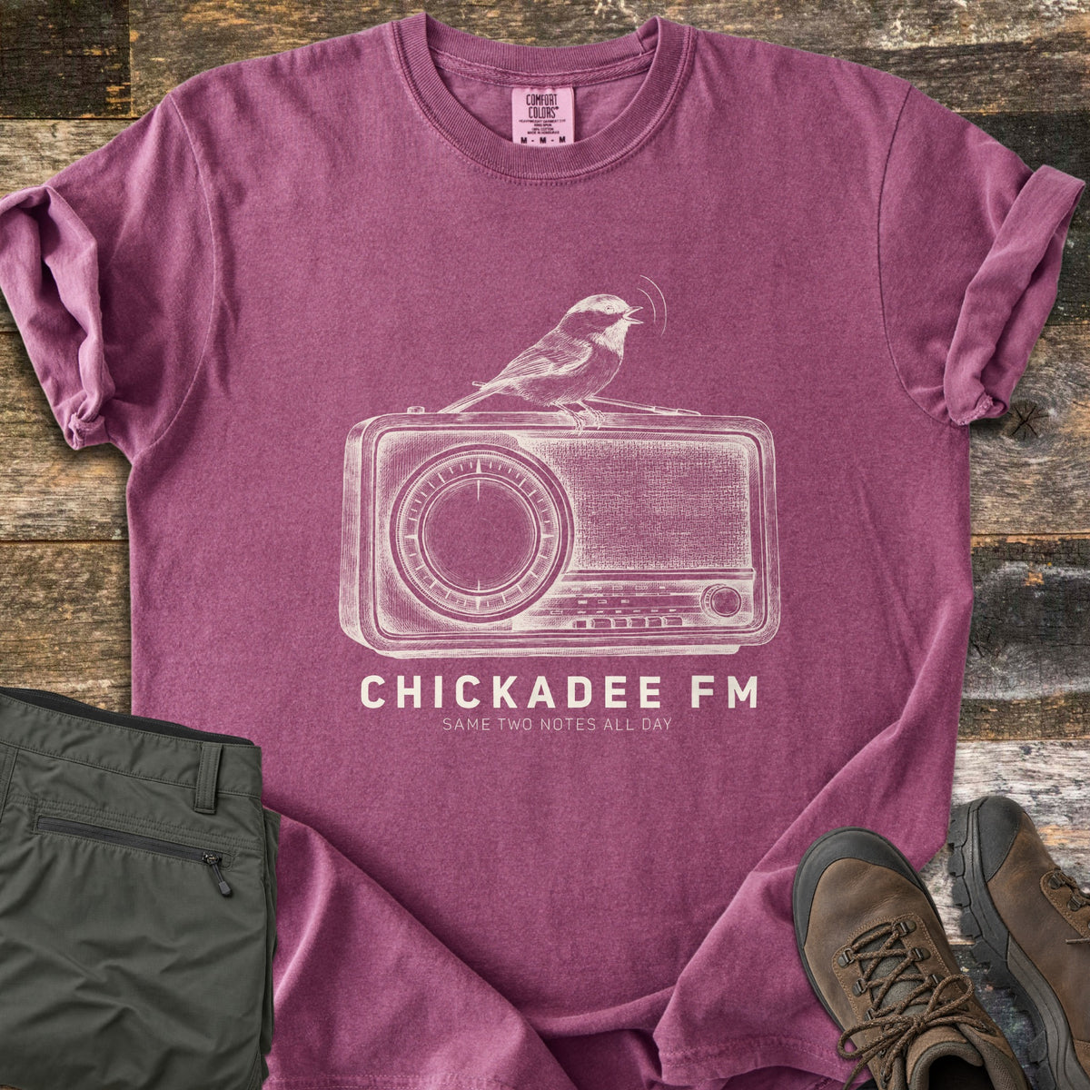 Chickadee FM Comfort Colors T-shirt in Berry, laid flat. A sketched chickadee singing atop a vintage radio, above the words CHICKADEE FM and Same Two Notes All Day in cream. Sleeves rolled, on weathered wood with folded green hiking pants and brown hiking boots.