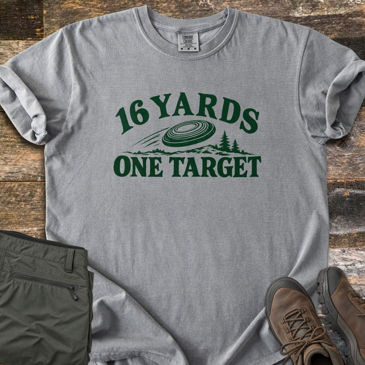 16 Yards One Target Comfort Colors T-shirt in Grey, laid flat. Dark green print of a clay target flying over a hilly treeline with pine trees, between the arched words 16 Yards and One Target. Olive cargo pants and brown hiking boots lie beside it on weathered wood planks.