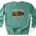 Trailside Table for Two Comfort Colors crewneck sweatshirt in Light Green, laid flat. An illustrated couple in caps sits on a plaid picnic blanket among pine trees, sharing food and mugs beside a side-by-side off-road vehicle, under the words Trailside and above Table for Two. Shown on a white surface.