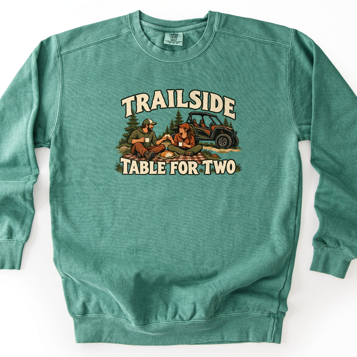 Trailside Table for Two Comfort Colors crewneck sweatshirt in Light Green, laid flat. An illustrated couple in caps sits on a plaid picnic blanket among pine trees, sharing food and mugs beside a side-by-side off-road vehicle, under the words Trailside and above Table for Two. Shown on a white surface.